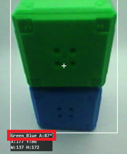 Ein Stapel aus zwei Würfeln, einer grün oben und einer blau unten, die von einem Sichtsystem verfolgt werden. Beide Würfel sind von einer weißen Umrandung umgeben, in der Mitte des grünen Würfels befindet sich ein weißes Kreuz. Die Beschriftung unten im Bild zeigt Grün_Blau A:87° und gibt damit die erkannten Farben und eine Winkelmessung an. Darunter sind die Koordinaten X:117, Y:186 mit den Abmessungen B:137, H:172 aufgeführt, die die Position und Größe der gestapelten Würfel im Rahmen darstellen.