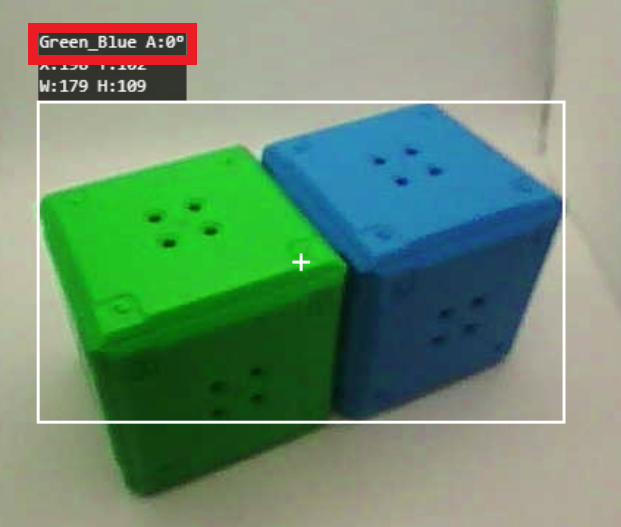 ลูกบาศก์ 2 ลูก สีเขียว 1 ลูก และสีน้ำเงิน 1 ลูก วางเคียงข้างกันและติดตามด้วยระบบการมองเห็น มีโครงร่างสีขาวล้อมรอบลูกบาศก์ทั้งสองลูก และมีกากบาทสีขาวอยู่ตรงกลาง ป้ายกำกับด้านบนซ้ายระบุเป็นสีเขียว_น้ำเงิน A:0° ซึ่งอ้างอิงถึงสีที่ตรวจพบและการวัดมุม ด้านล่างนั้น พิกัดจะแสดงเป็น X:150, Y:102 โดยมีมิติ W:179, H:109 ซึ่งแสดงตำแหน่งและขนาดของลูกบาศก์ภายในกรอบ