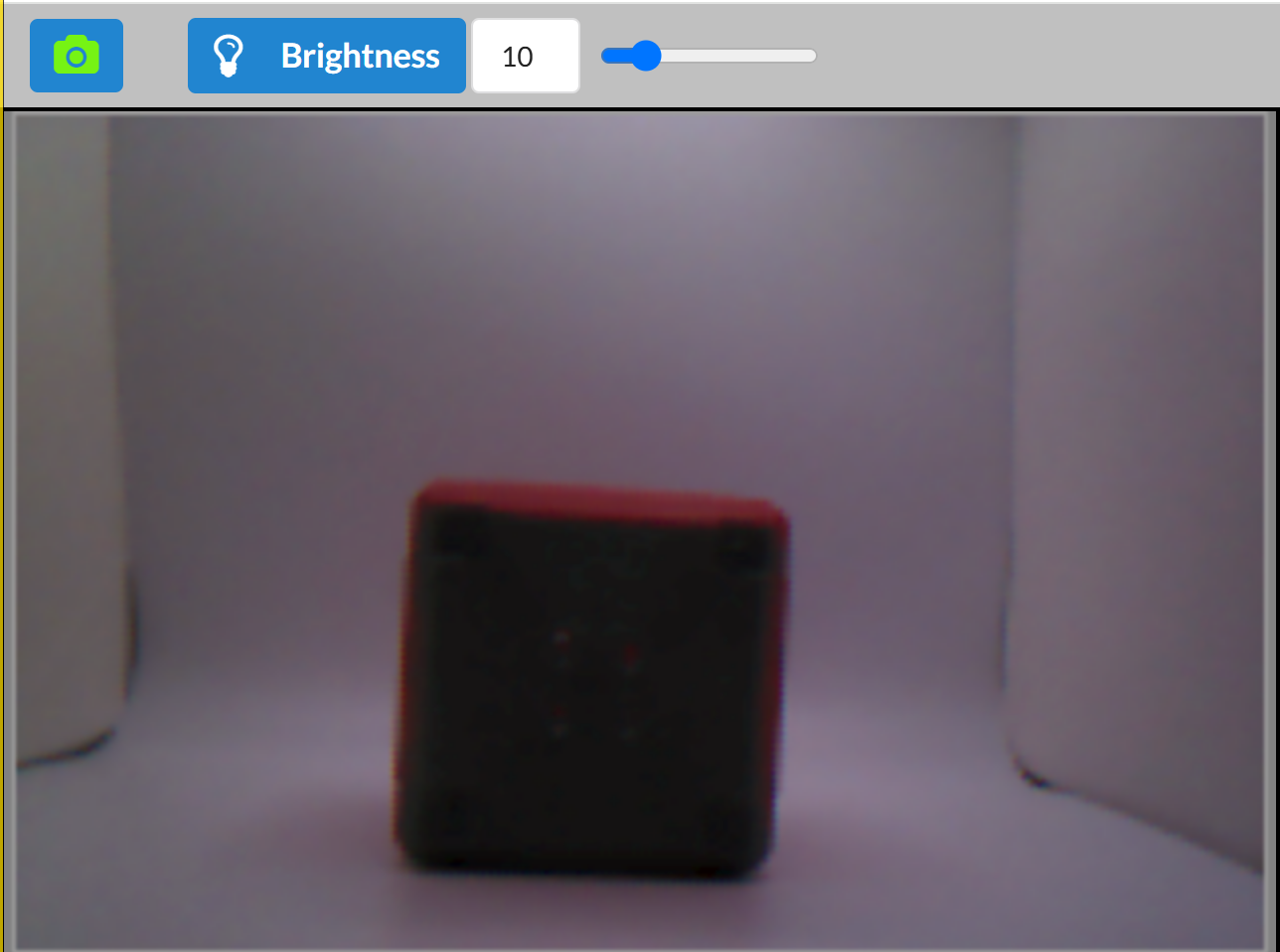 The Vision Sensor preview showing a red cube with brightness set to 10. The scene appears underexposed but more visible than the previous image. The cube is centered in a curved white backdrop and is tilted slightly to the right.