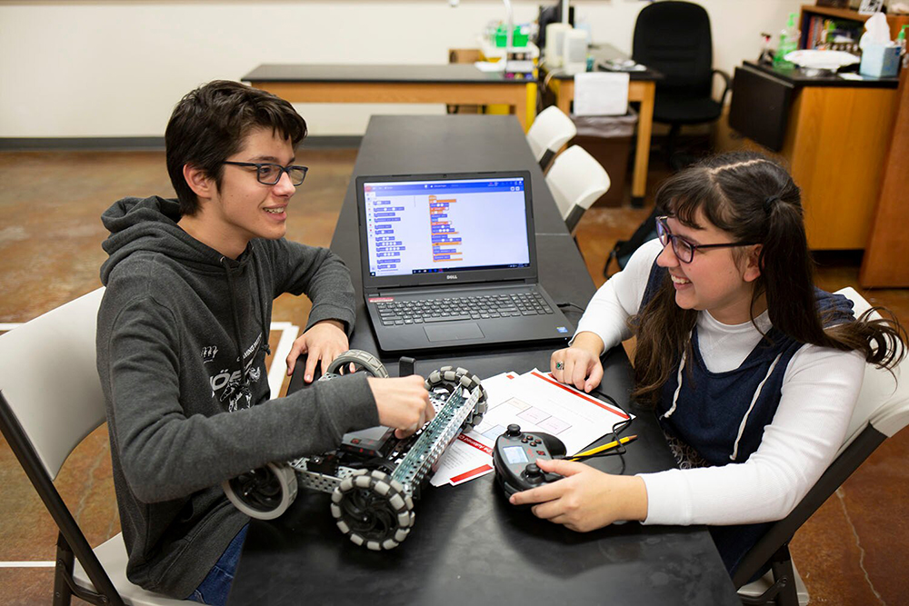 Zēns un meitene strādā kopā ar robotu VEX V5, demonstrējot sadarbību STEM izglītībā. Bērni ir iesaistīti praktiskā apmācībā, uzsverot robotikas nozīmi izglītības iestādēs.