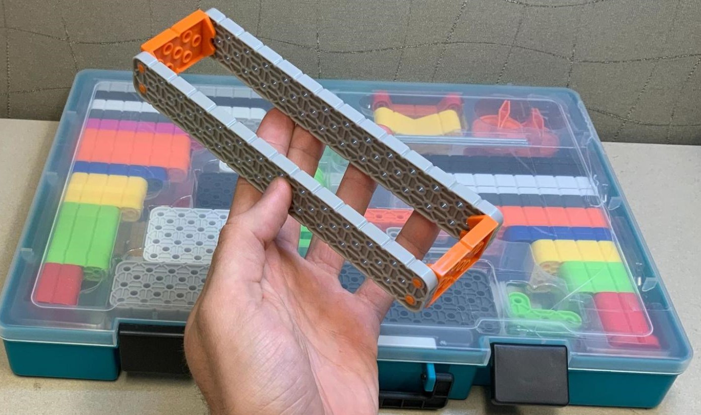 Eine Hand, die die beiden großen Balkenteile hält, die mithilfe von zwei orangefarbenen Verbindungsstücken verbunden sind, und zwar in einer ähnlichen Ausrichtung wie im Beispiel der Bauanleitung.