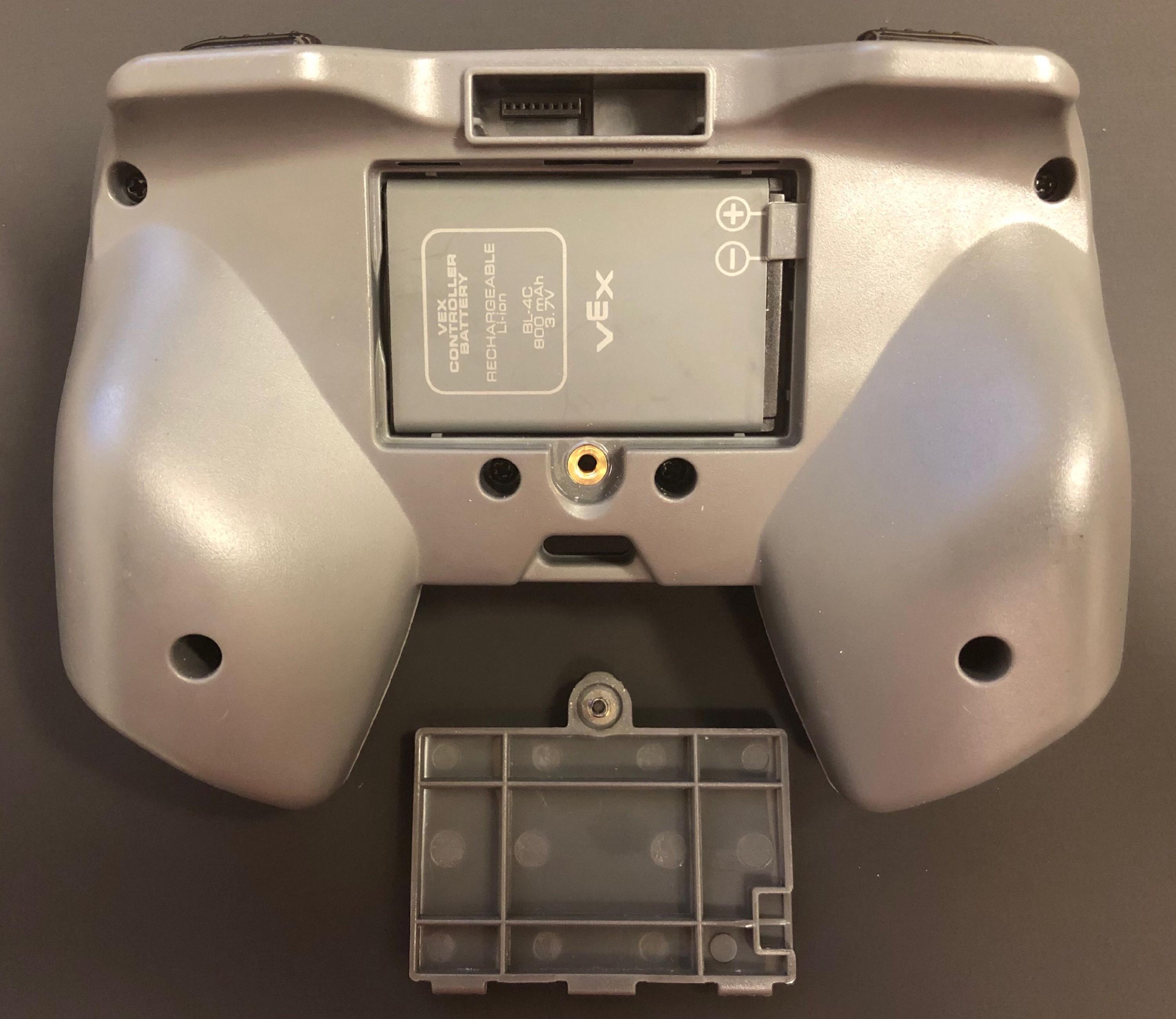 Controlador con la puerta de la batería abierta y una batería nueva colocada en ella. La batería tiene íconos positivos y negativos que deben estar alineados con los del controlador. En esta imagen, los íconos de la batería están en la esquina superior derecha de la ranura de la batería.