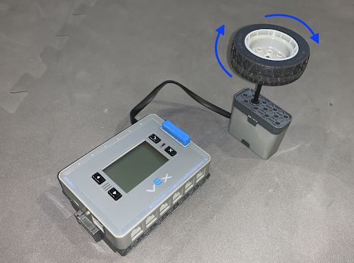 Un cerveau IQ avec une batterie est placé sur une table. Un moteur intelligent est connecté à l’un de ses ports intelligents. Un arbre moteur fixé à une pièce de roue est connecté au moteur intelligent. Les flèches indiquent que la pièce de la roue doit être tournée rapidement.