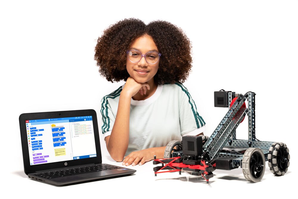 Một học sinh đang ngồi gần V5 Clawbot và một máy tính xách tay có dự án VEXcode V5 Blocks hiển thị trên màn hình.