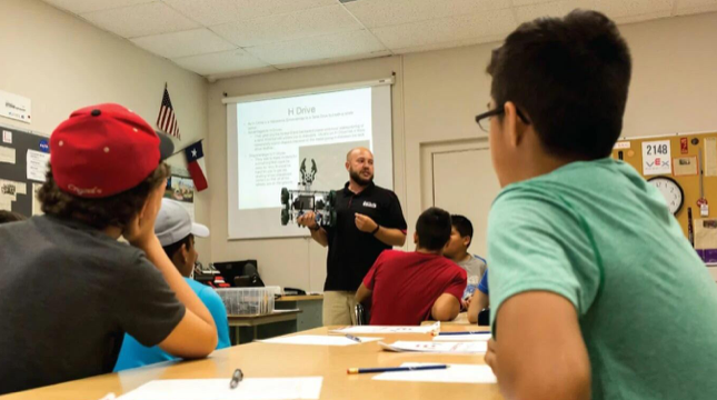 Um grupo de alunos participou numa atividade prática de aprendizagem STEM com o VEX V5. Um professor está de pé, em frente à sala de aula, segurando um robô V5.