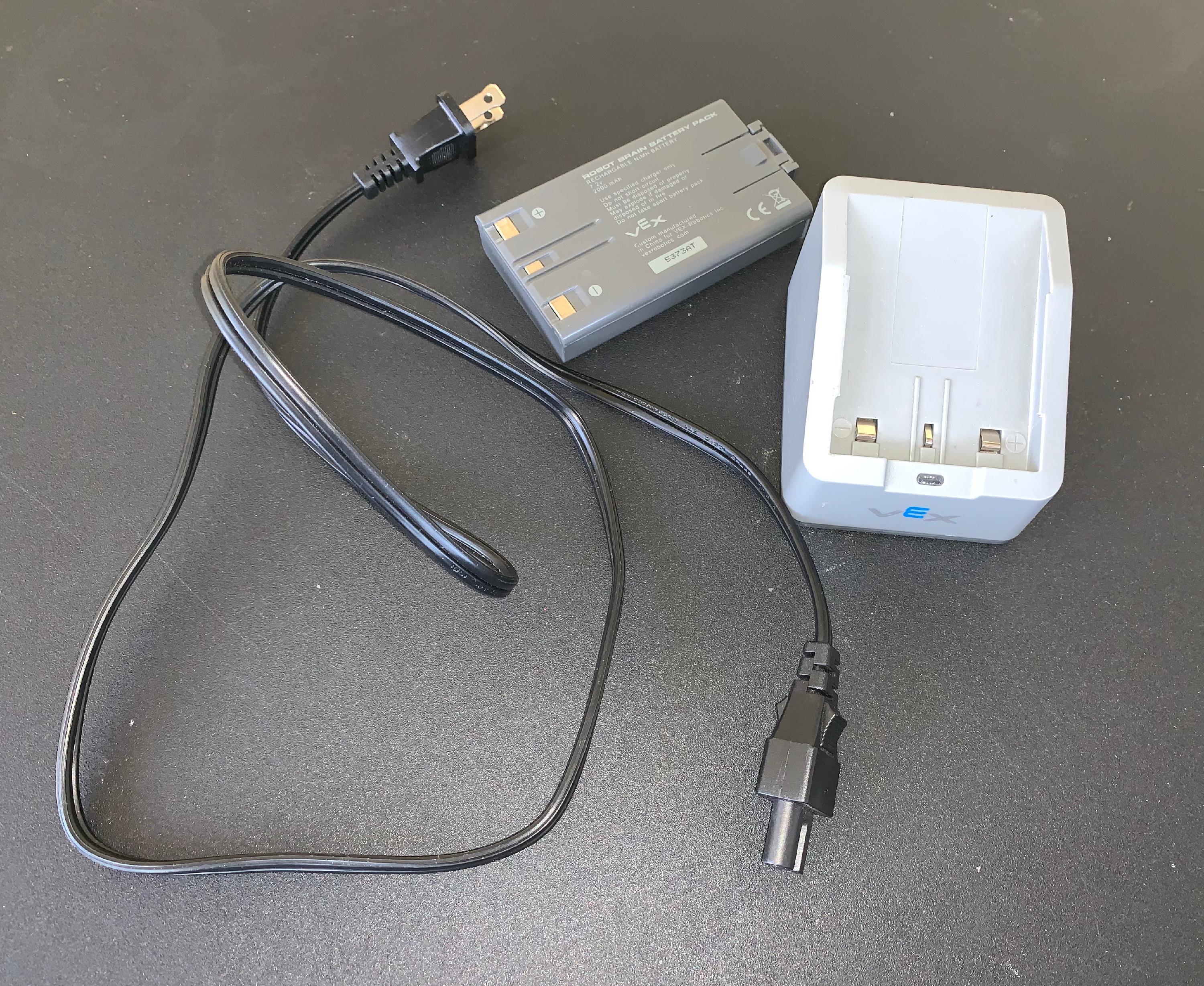 Ett robotbatteri, en robotbatteriladdare och en batteriladdarens nätsladd placerade tillsammans på ett bord.