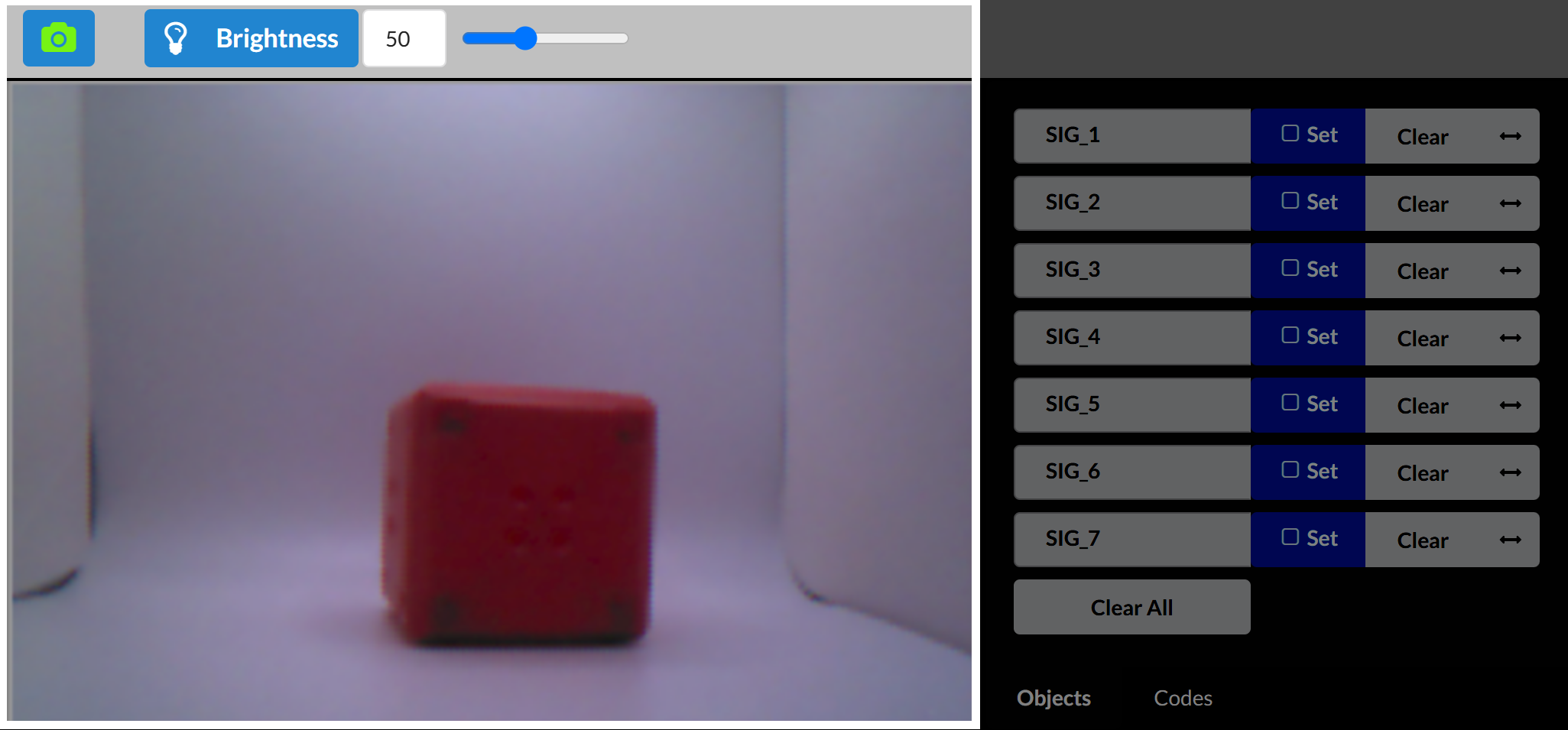 Il feed di anteprima in tempo reale di Vision Sensor Utility mostra un cubo rosso centrato in uno spazio di calibrazione bianco. L'interfaccia mostra in alto un cursore Luminosità impostato su 50 e sul lato destro i controlli di configurazione per SIG_1 fino a SIG_7. Il pulsante Cancella tutto appare sotto gli slot della firma.