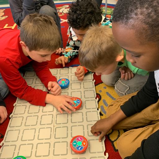 A group of students sits on the floor around a 123 Field touch coding and watching the robots move.