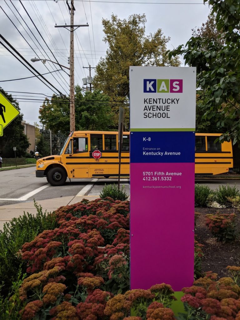 A school sign on a street corner reads KAS Kentucky Avenue School in front of a school bus.
