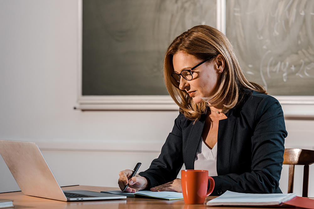 A female teacher sits making planning notes at a table in front of an open laptop.