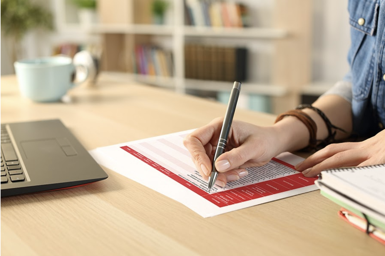 A teacher writes on a V5 rubric at a desk beside an open laptop.