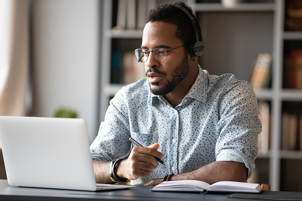 A teacher sits at a desk planning on paper with an open laptop in front of him.