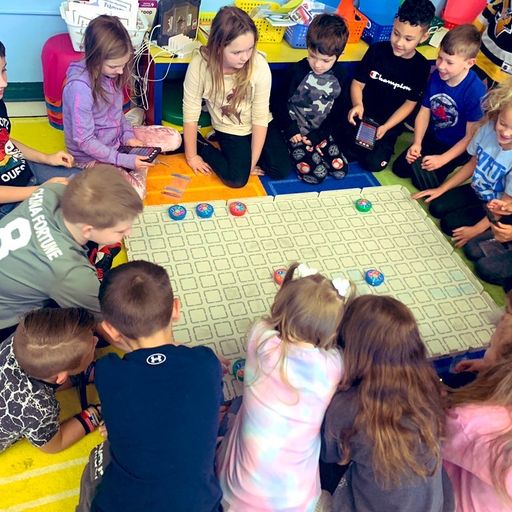 A class of students sits around a large 123 Field watching several robots move around to solve a challenge.