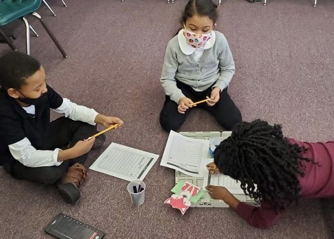 Three students sit on the floor around a 123 Field, with a robot, red wolf, and Coder as they write their project plan in a worksheet.