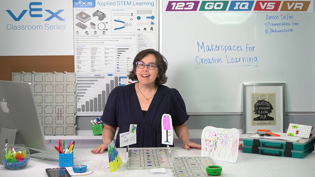 A woman stands behind a table with VEX 123 and VEX GO projects in front of her, and the board reads Makerspaces for Creative Learning.