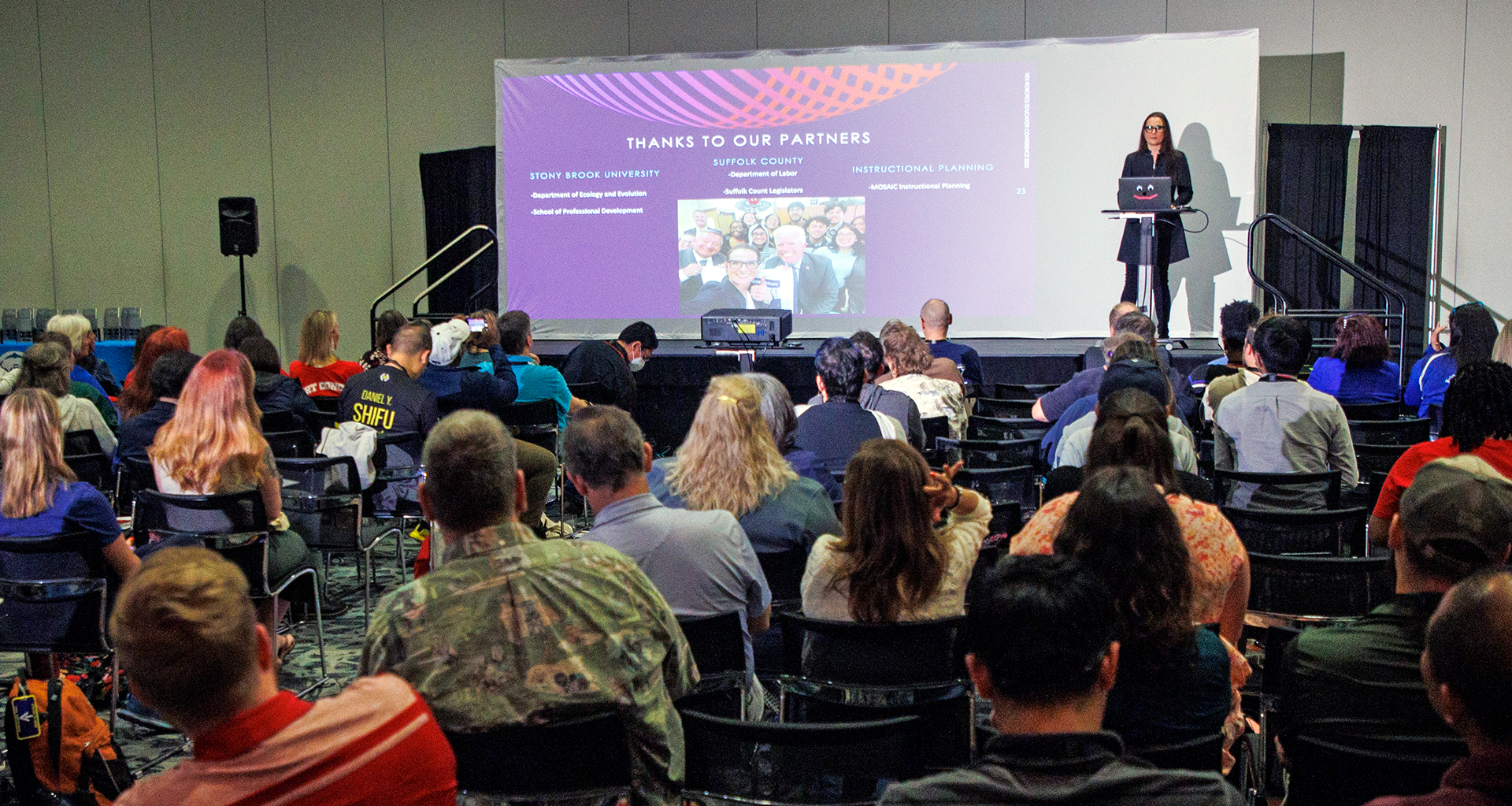 A woman is pesenting in a large conference room at the VEX Robotics Educators Conference, in front of a live audience.