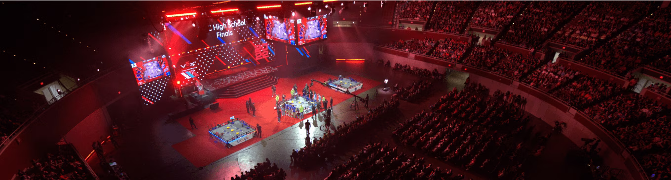 VEX Worlds Competition event, showing students gathered around a VEX field in front of a large audience.