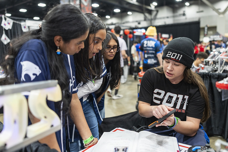 4 kids talking with one another. An engineering notebook can be seen in the center with the girl on the right pointing at a tablet. The other students are engaged to see how they documented their process.