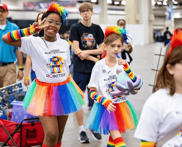 2 girls wearing robotics team t-shirts pull their VEX IQ robot in a wagon at a competition.