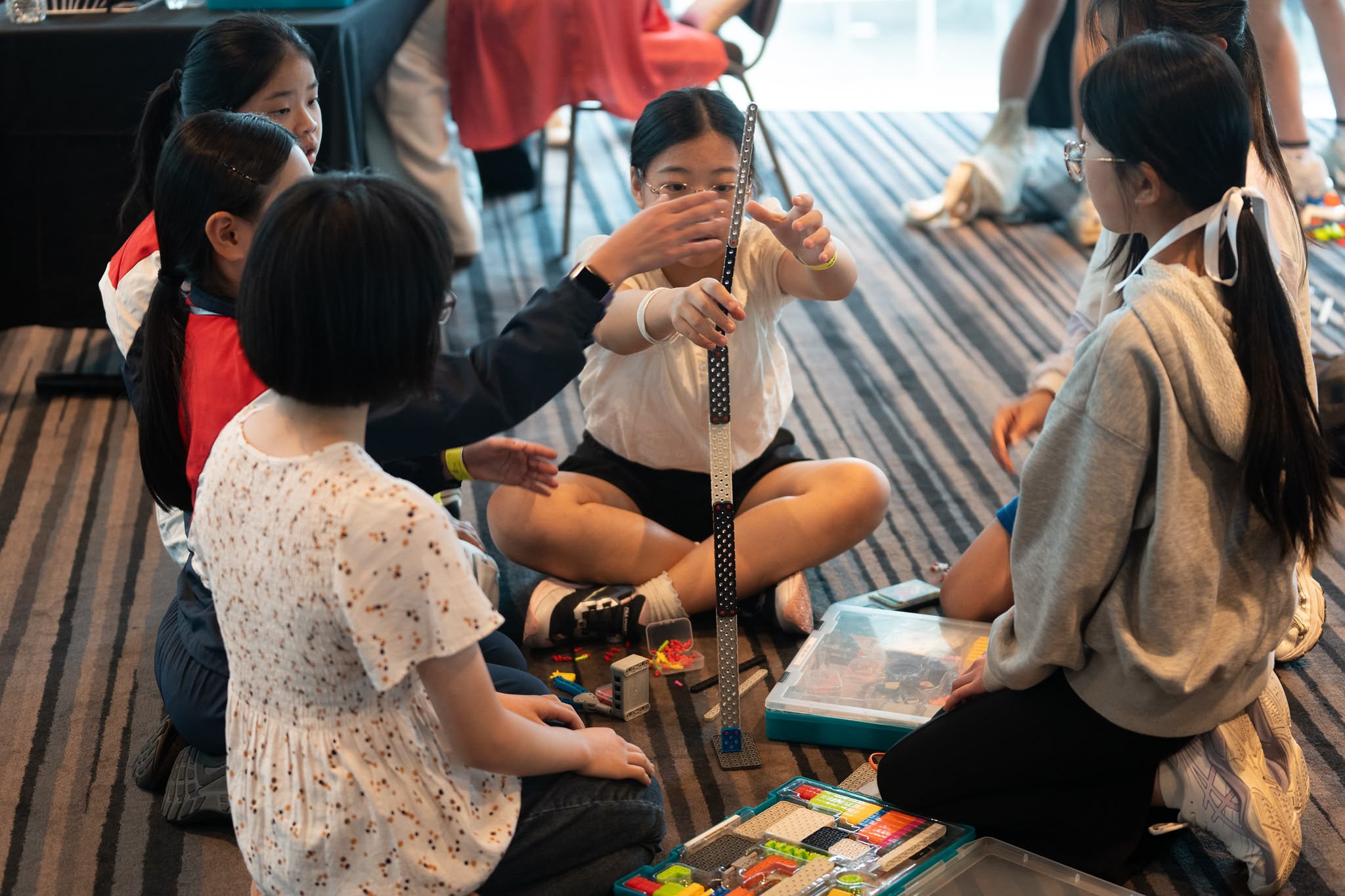 5 girls seated on a rug building a tower with VEX GO. Two girls cooperate to hold the tower in place.