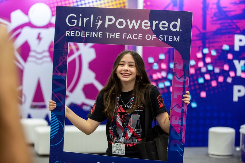 A smiling female student stands holding a Girl Powered promotional frame around her face at an event.