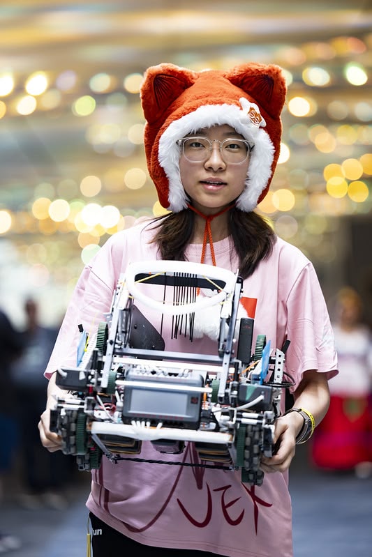 A female student in a team shirt and a fox hat carries a metal robot at a competition event.