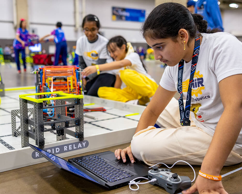 Two girls position a robot on a practice field, while the third works at a laptop to adjust the controller code.