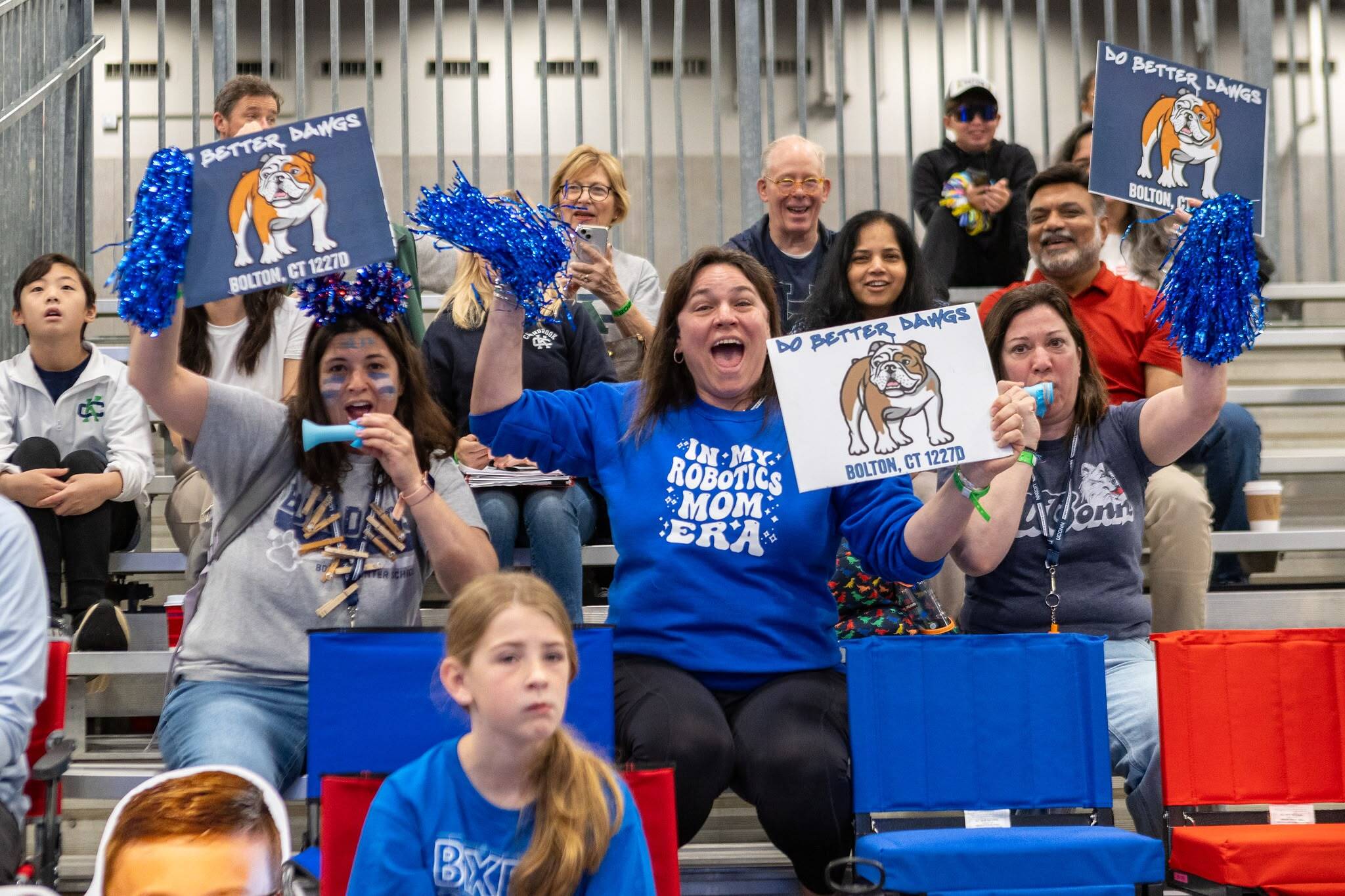 أفراد الجمهور الجالسون في المدرجات يشاهدون المباراة وهم يهتفون حاملين كرات التشجيع ولافتات عليها شعار الفريق. في المنتصف، تهتف امرأة وهي ترتدي قميصاً كُتب عليه "في عصر أمي في مجال الروبوتات".