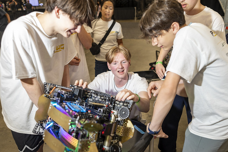 Para siswa mengelilingi robot untuk memperbaikinya sementara seorang pelatih mengawasi dari belakang. Mereka mengamati dan membiarkan para siswa mengarahkan perubahan apa pun pada robot tersebut.