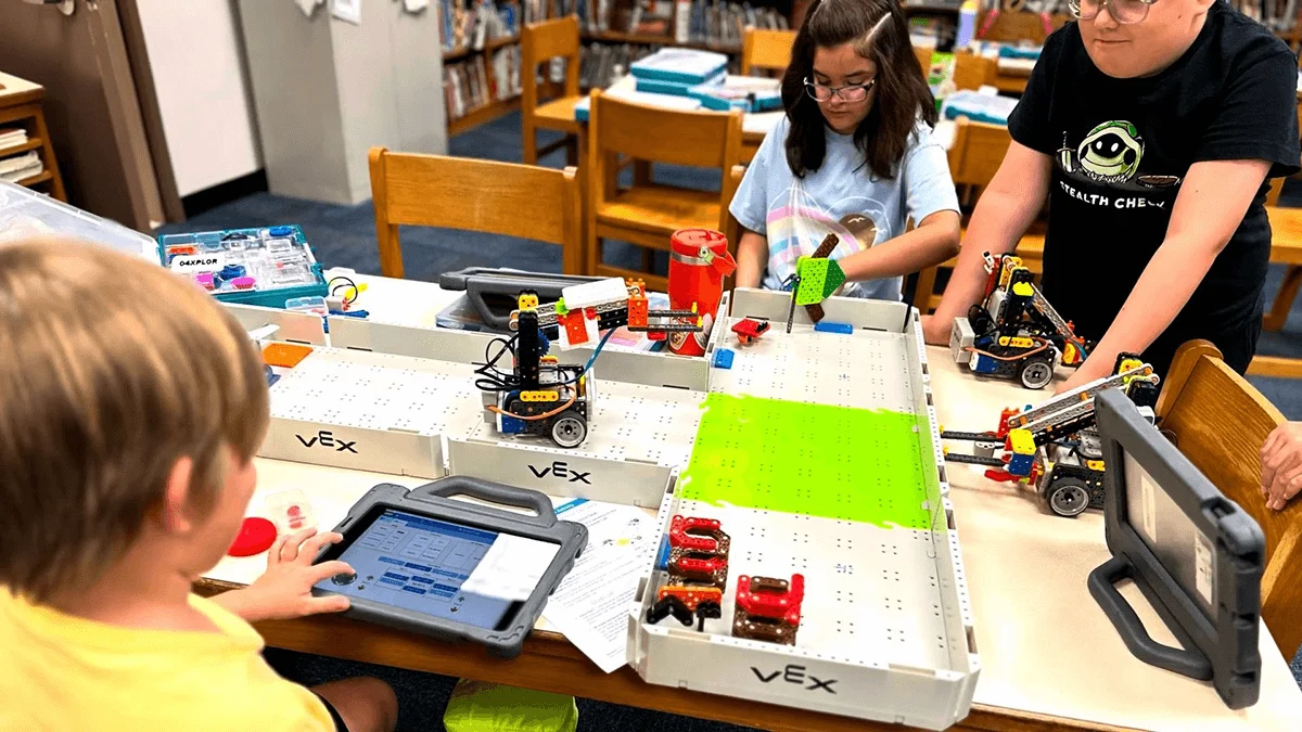 Studenti strādā bibliotēkā ap galdu, ap kuru atrodas VEX GO laukums un sacensību roboti. Vienam studentam ir iPad, un viņš kodē robotu, bet pārējie divi pozicionē robotu un piedāvā idejas.